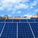 Rows of blue solar panels installed on a rooftop under a bright blue sky with scattered clouds and traditional-style buildings in the background.